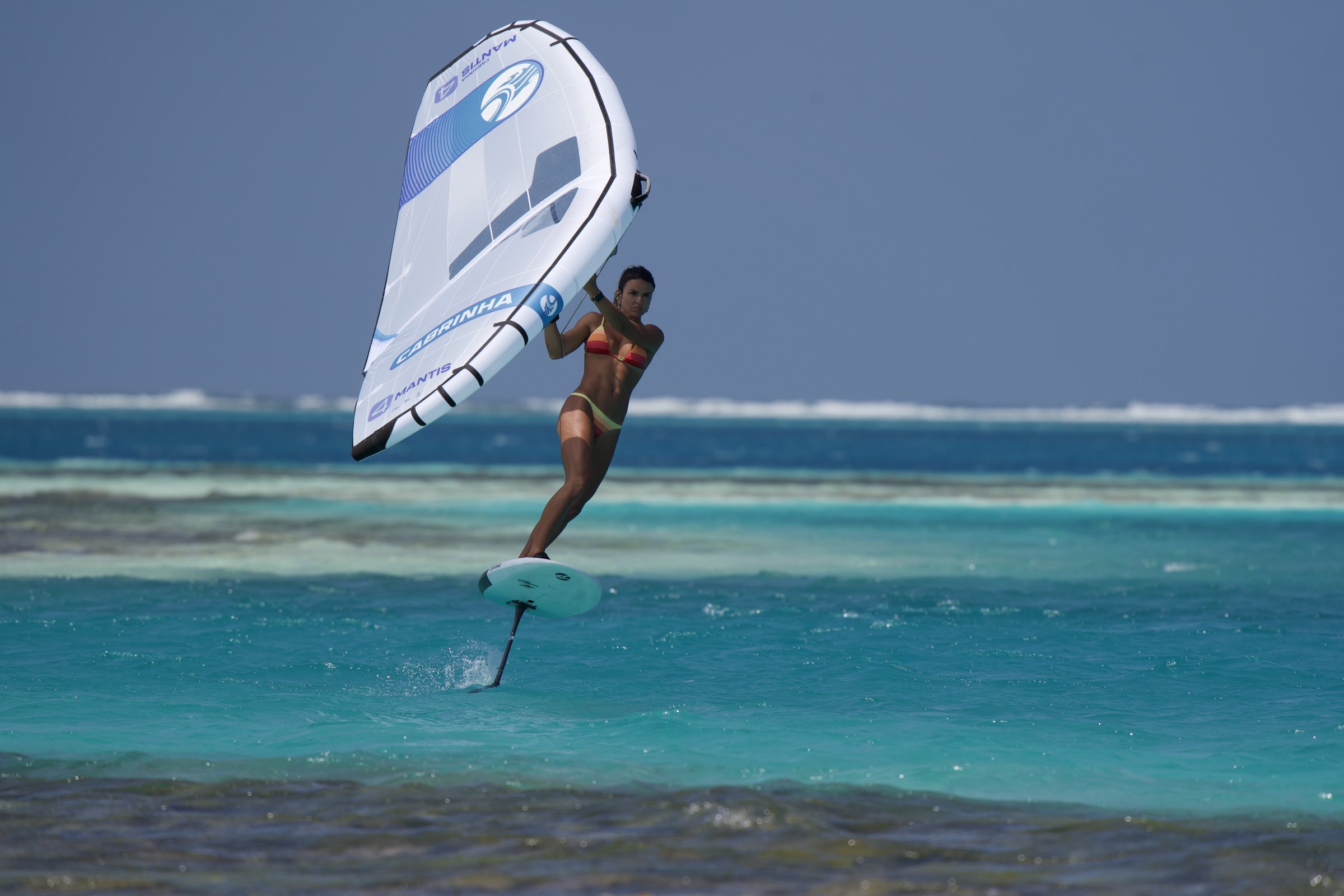 Manu La Porta voando de wingfoil sobre águas turquesa, com o wing branco e azul aberto